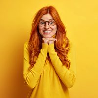 Joyful surprised redhead young woman keeps hands under chin shows white teeth feels glad listens with great interest interlocutor dressed in casual jumper isolated over vivid yellow background